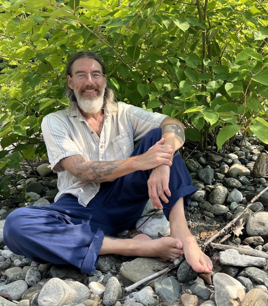 Timothy Lee Scott, herbalist, author and health guide, smiling as he sits barefoot on river stones beneath Japanese knotweed